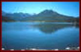 This is a photo of Silver Jack Reservoir. This view shows a calm, very blue lake with distant vegetation covered jagged mountains. At the bottom of the photo is driftwood held behind a floating barrier. Select for a larger photo.